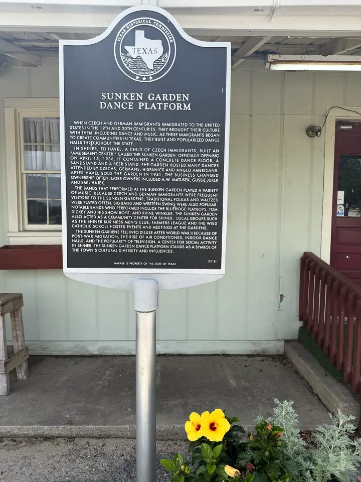 The Sunken Garden Dance Platform Texas historical marker in front of the historic Sunken Gardens building, with yellow hibiscus flowers below.