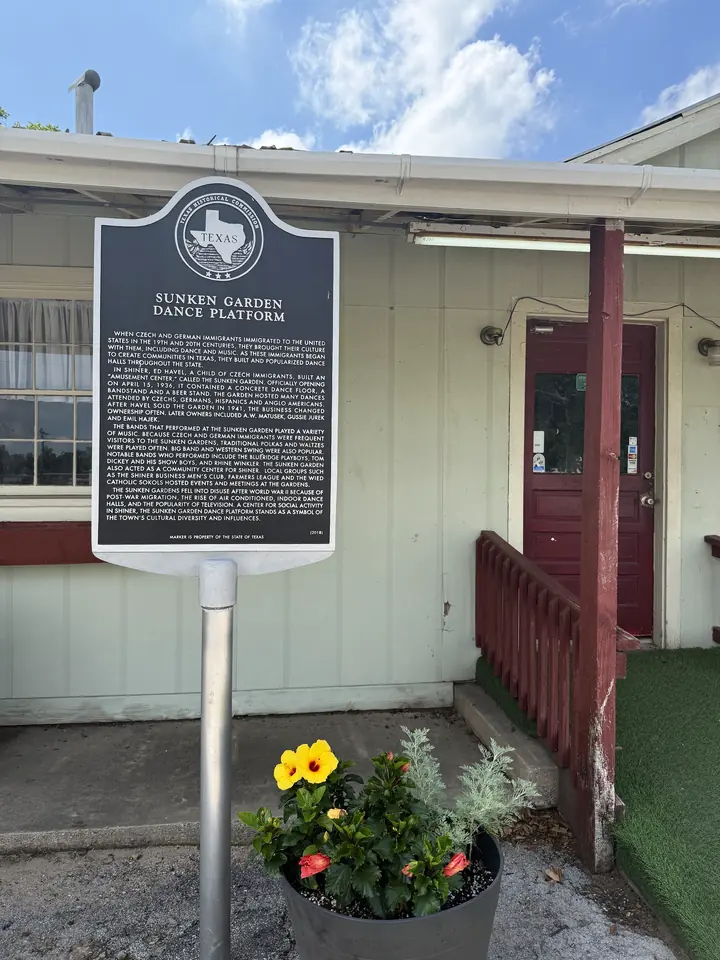 The Sunken Garden Dance Platform marker, hibiscus flowers, a planter, and the historic red-door building entrance.