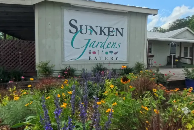 The Sunken Gardens sign behind a bright front flower bed with purple salvia and orange blooms.
