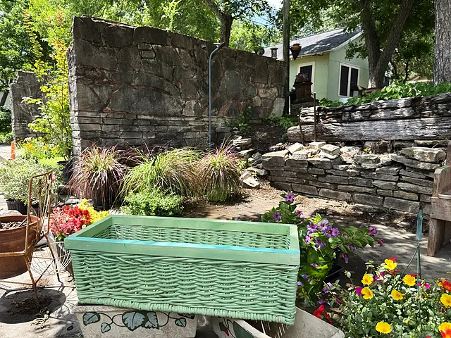 A green woven basket-style container on the stone terrace with grasses and flowers behind it.