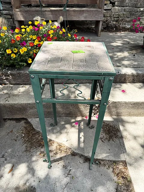 A green metal plant stand on the patio in front of flowers and stone steps.
