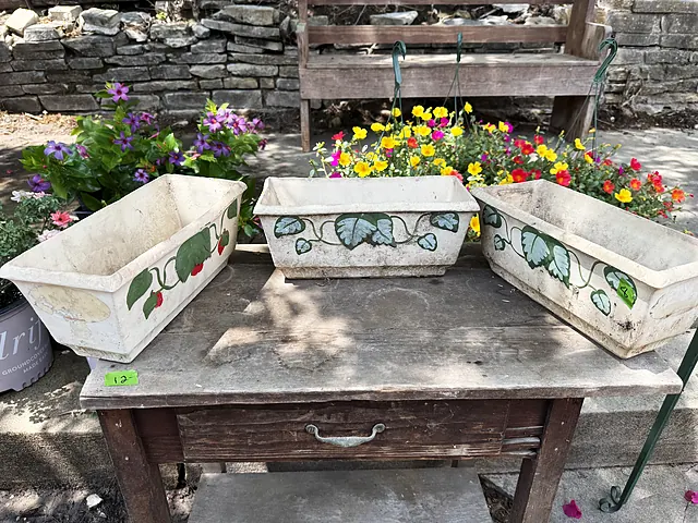 Painted rectangular planters on a weathered worktable with bright flowers behind them.