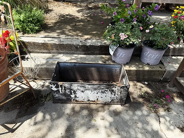 A rectangular metal planter staged near stone steps and potted flowers.