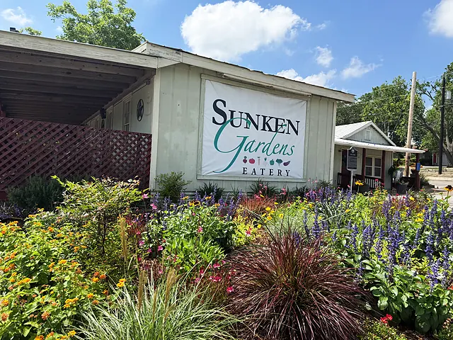 The Sunken Gardens Eatery sign framed by flower beds, grasses, and the shaded porch.