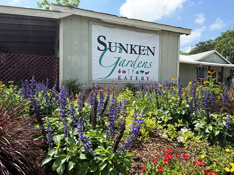 The Sunken Gardens Eatery sign behind purple salvia, red blooms, and summer garden color.