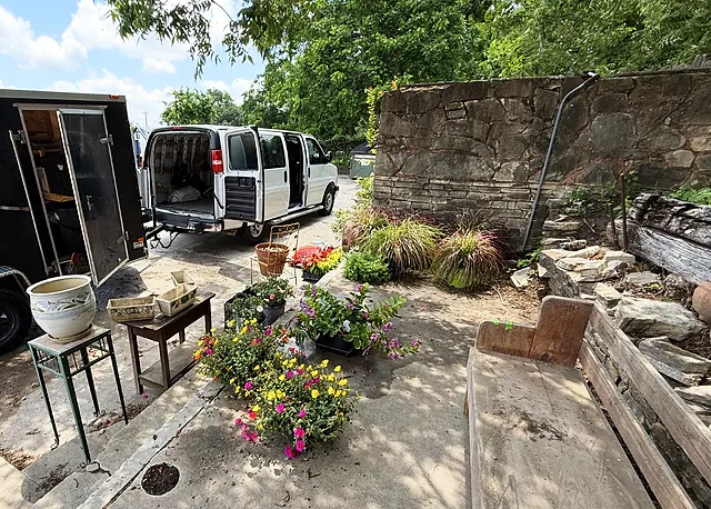A wide setup view with a trailer, work van, plants, containers, and old stone walls.
