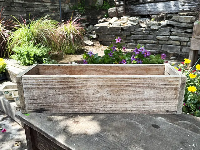 A weathered wooden planter box on a table with flowers and stonework behind it.